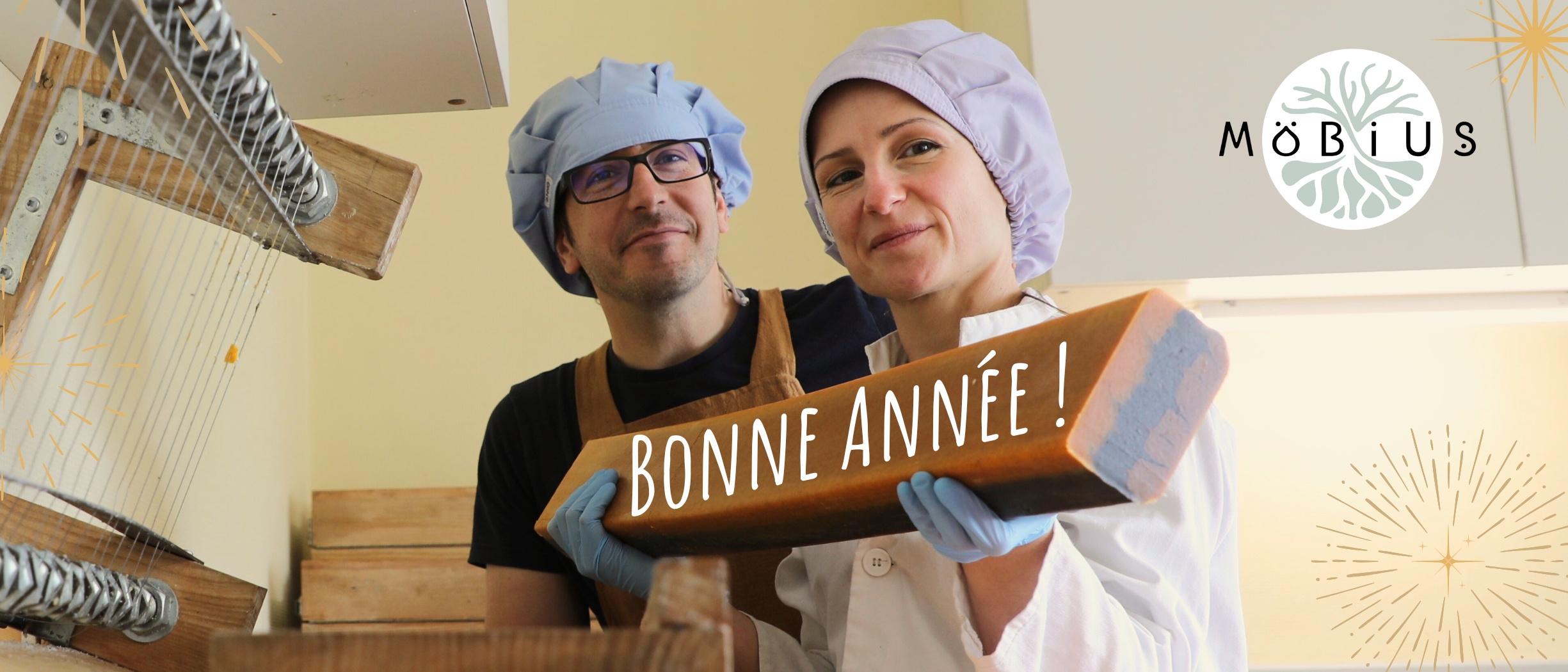 Virginie et Hubert dans leur laboratoire tenant un gros pain de savon "Bonne Mine" avec le message "Bonne année !" ajouté numériquement, près d'une guitare à découper le savon.