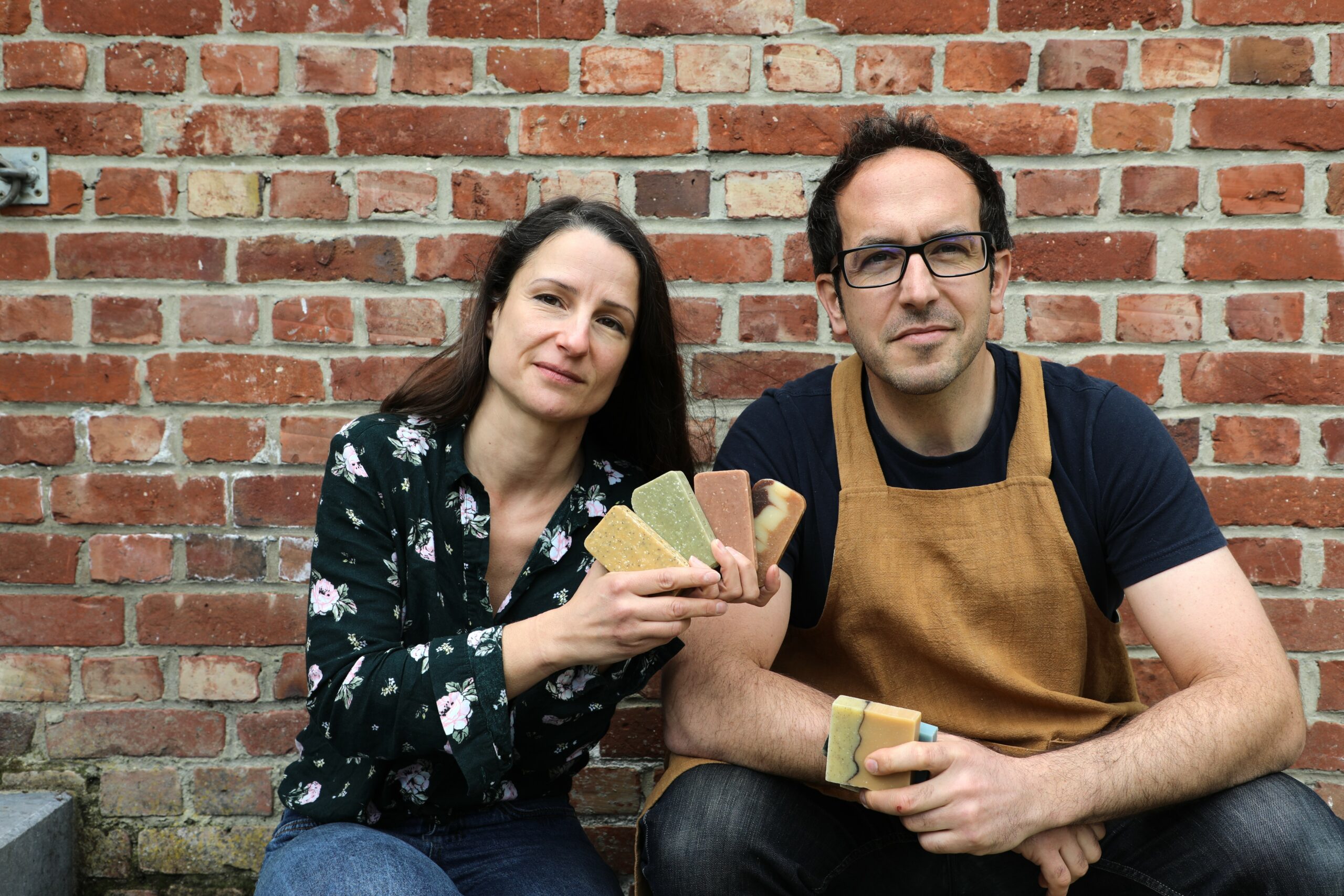 Virginie et Hubert devant un mur de briques, tenant quelques savons MöBiUS dans leurs mains.