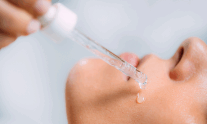 Gros plan sur un visage de profil avec une peau lumineuse. Une main tient une pipette laissant couler un sérum clair sur la joue, illustrant un soin hydratant et nourrissant pour la peau.