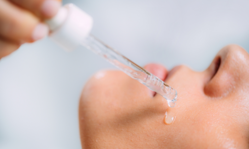 Gros plan sur un visage de profil avec une peau lumineuse. Une main tient une pipette laissant couler un sérum clair sur la joue, illustrant un soin hydratant et nourrissant pour la peau.
