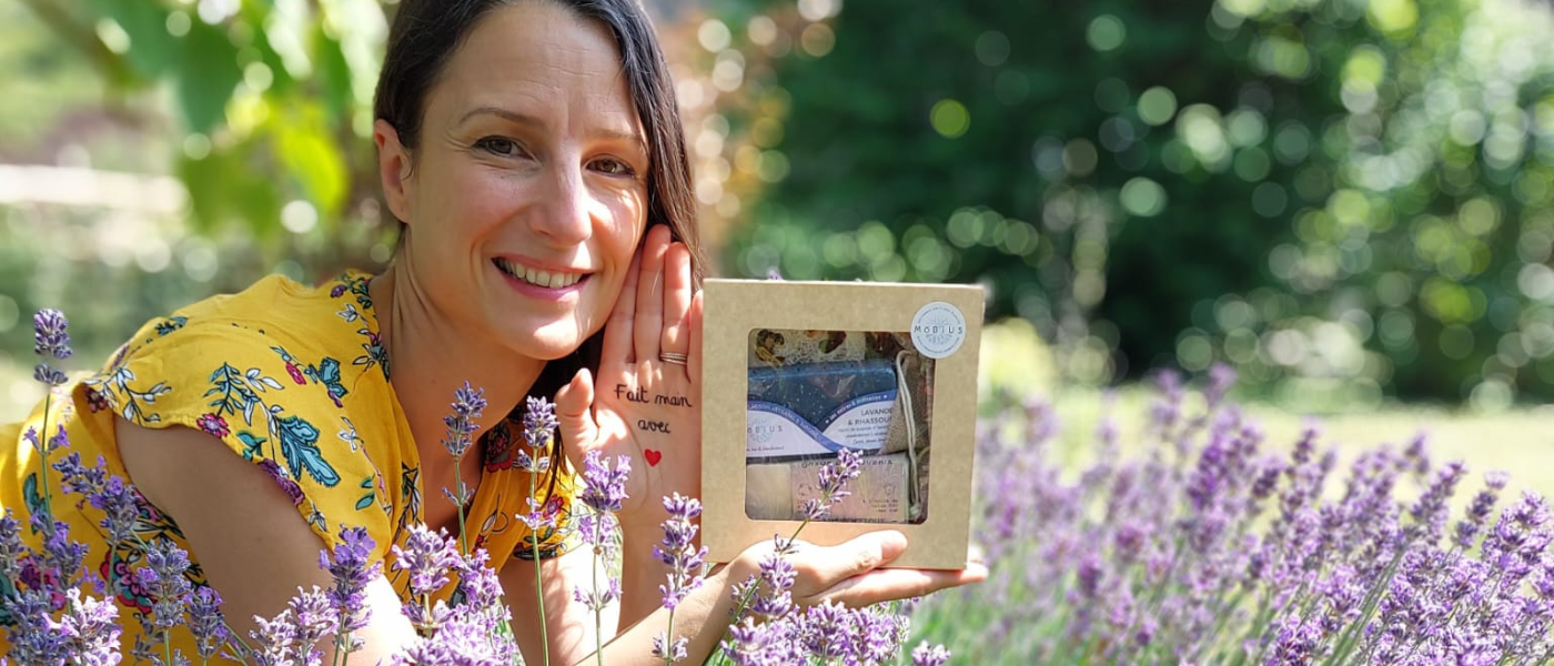 Virginie tenant un coffret cadeau MöBiUS avec savon lavande & rhassoul, au milieu d'un champ de lavande, avec la main tendue portant l'inscription "fait main avec ❤️" et arrière-plan naturel flou.