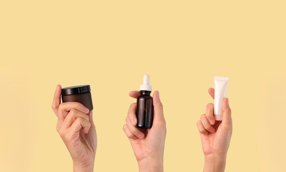 Trois mains féminines tenant un pot ambré, un flacon pipette et un tube de crème sur fond jaune soleil – illustration des trois textures cosmétiques : baume, sérum et crème.
