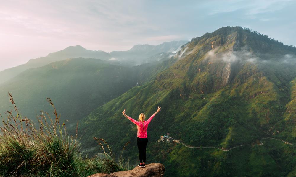Femme en tenue de sport vue de dos, debout sur une avancée rocheuse, les bras levés en V face à un paysage de montagne