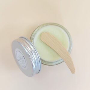 Spatule cosmétique en bois de bambou posée sur un pot de baume déodorant MöBiUS, pot ouvert avec couvercle à côté, fond orangé pastel
