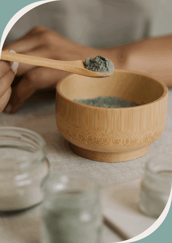 Pot en bois rempli d’argile verte en poudre, avec une main floue tenant une cuillère en bois, et des pots en verre flous au premier plan