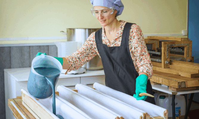 Virginie en tenue de laboratoire verse la pâte du savon saponifiée à froid lavande & rhassoul dans les moules, avec plusieurs moules au premier plan et casseroles en arrière-plan.
