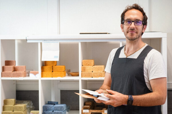 Hubert souriant tenant une enveloppe de la poste devant l’étagère de logistique avec savons saponifiés à froid, shampoings et déodorants prêts à être expédiés.
