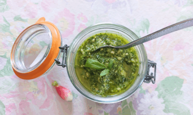 Petit bocal en verre ouvert rempli de pesto de fanes de radis, avec une cuillère enfoncée dedans, posé sur une nappe pastel fleurie.