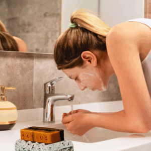 Femme rinçant son visage nettoyé au savon saponifié à froid ; savon saponifié à froid Bonne Mine posé sur un porte-savon près du lavabo.