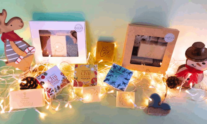 Coffrets cadeaux et savons artisanaux MöBiUS dans leurs boîtes festives, présentés pour Noël sur fond vert avec guirlande lumineuse.