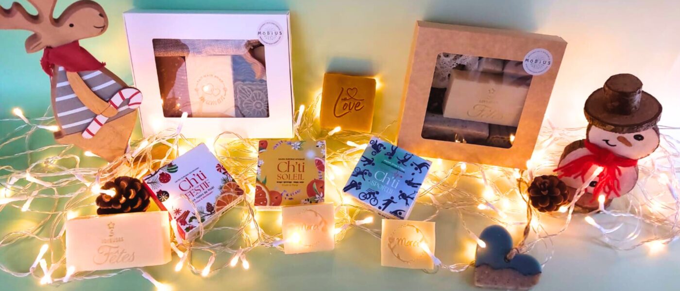 Coffrets cadeaux et savons fabriqué à Lille MöBiUS dans leurs boîtes festives, présentés pour Noël sur fond vert avec guirlande lumineuse.