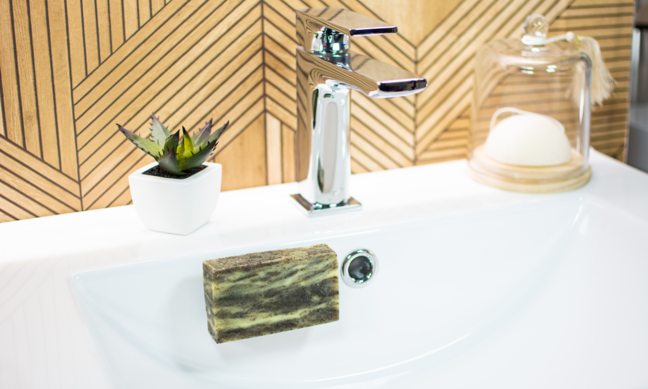 Savon exfoliant naturel au marc de café en lévitation dans un lavabo blanc, avec éponge de konjac sous cloche en verre et décoration végétale